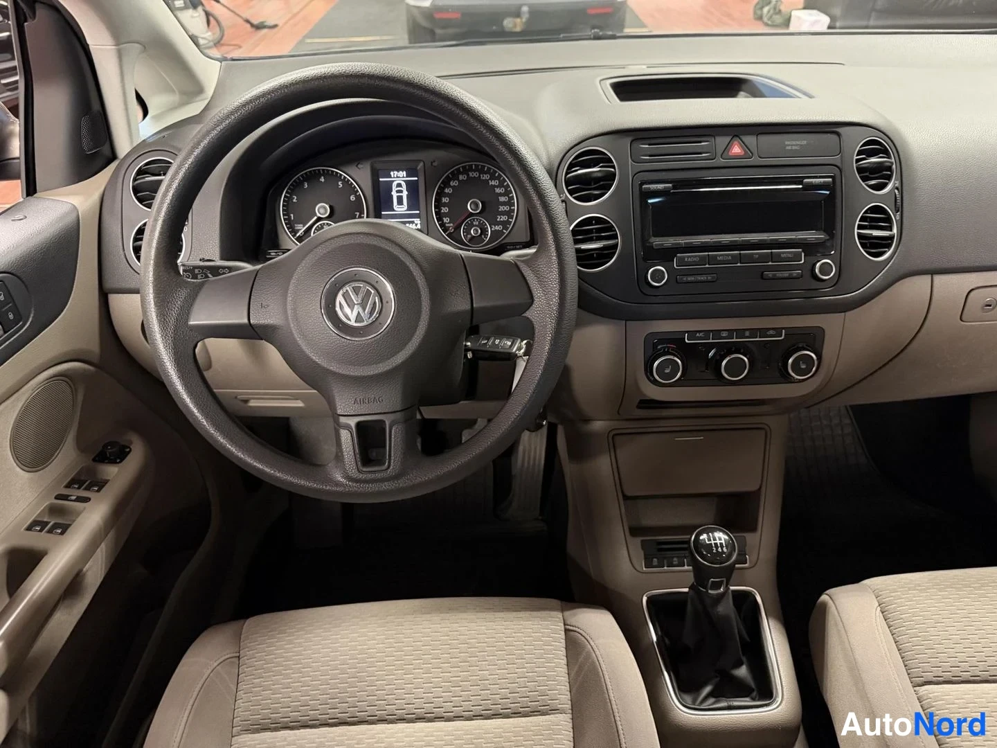 Edestä: Auton ohjaamo: VW-merkkinen ohjauspyörä, analogiset mittarit, radiolaite ja manuaalivaihteisto, kankaat istuimet etualalla.