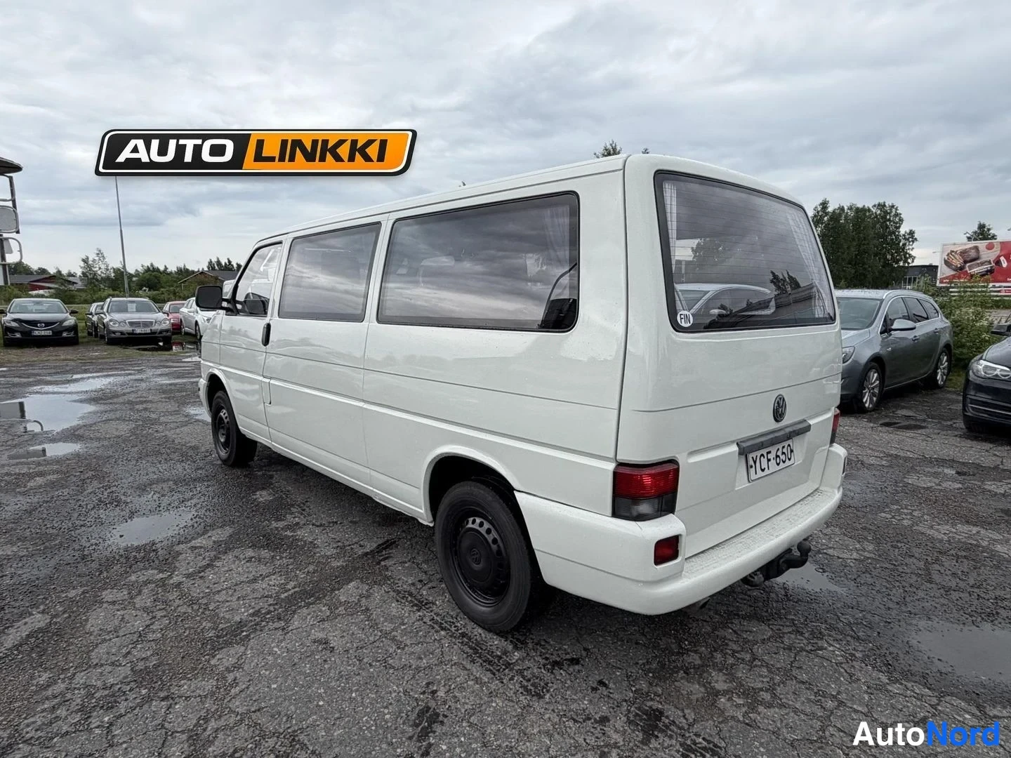 Takaviisto: Valkoinen Volkswagen Caravelle takaa kolmannelta sivulta, perä ja vasen kylki näkyvät rekisterikilven kanssa.
