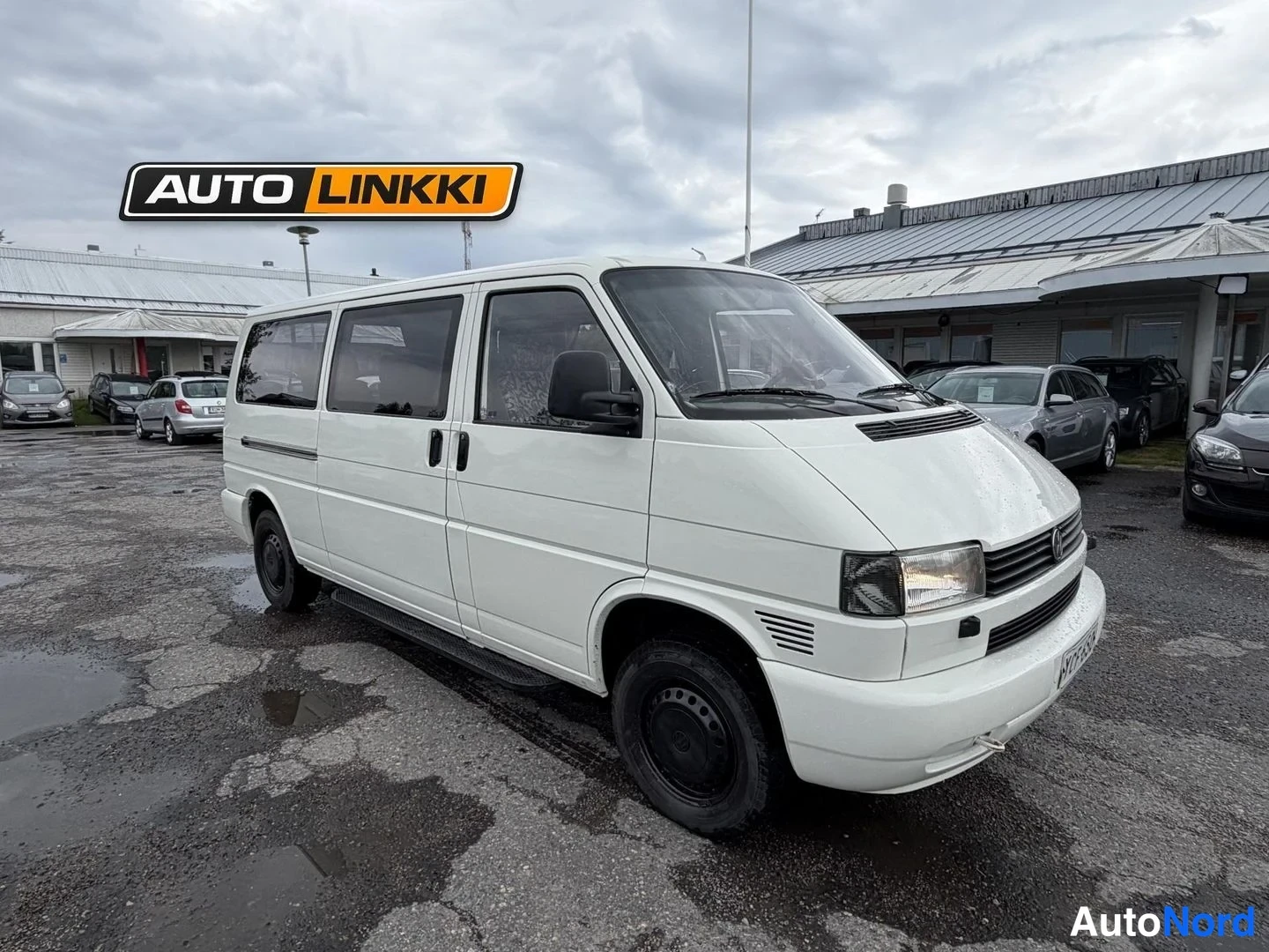 Etuviisto, nokka oikealle: Valkoinen pitkä pakettiauto etu- ja sivukulmasta kuvattuna autokorin pihalla, keula osoittaa oikealle.