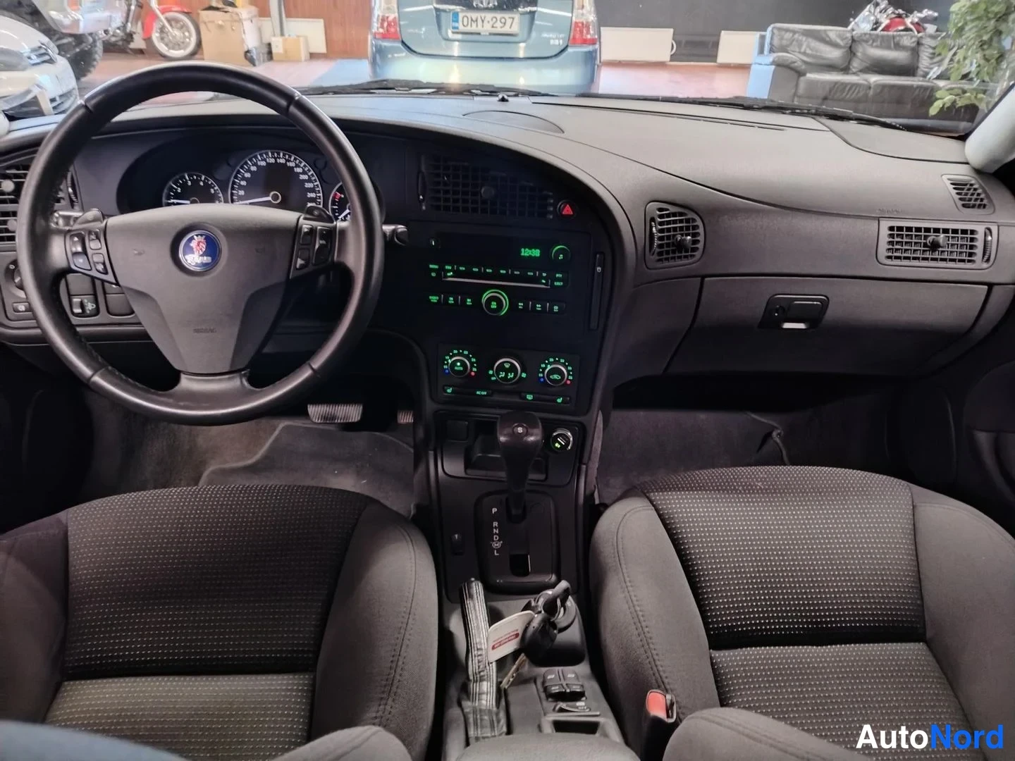 Muu: Saab 9-5:n ohjaamo näkyy tuulilasin läpi: musta kojelauta, vihreä valaistu radio, automaattivaihteen valitsin P-R-N-D-asennossa ja kaksi etuistuinta.