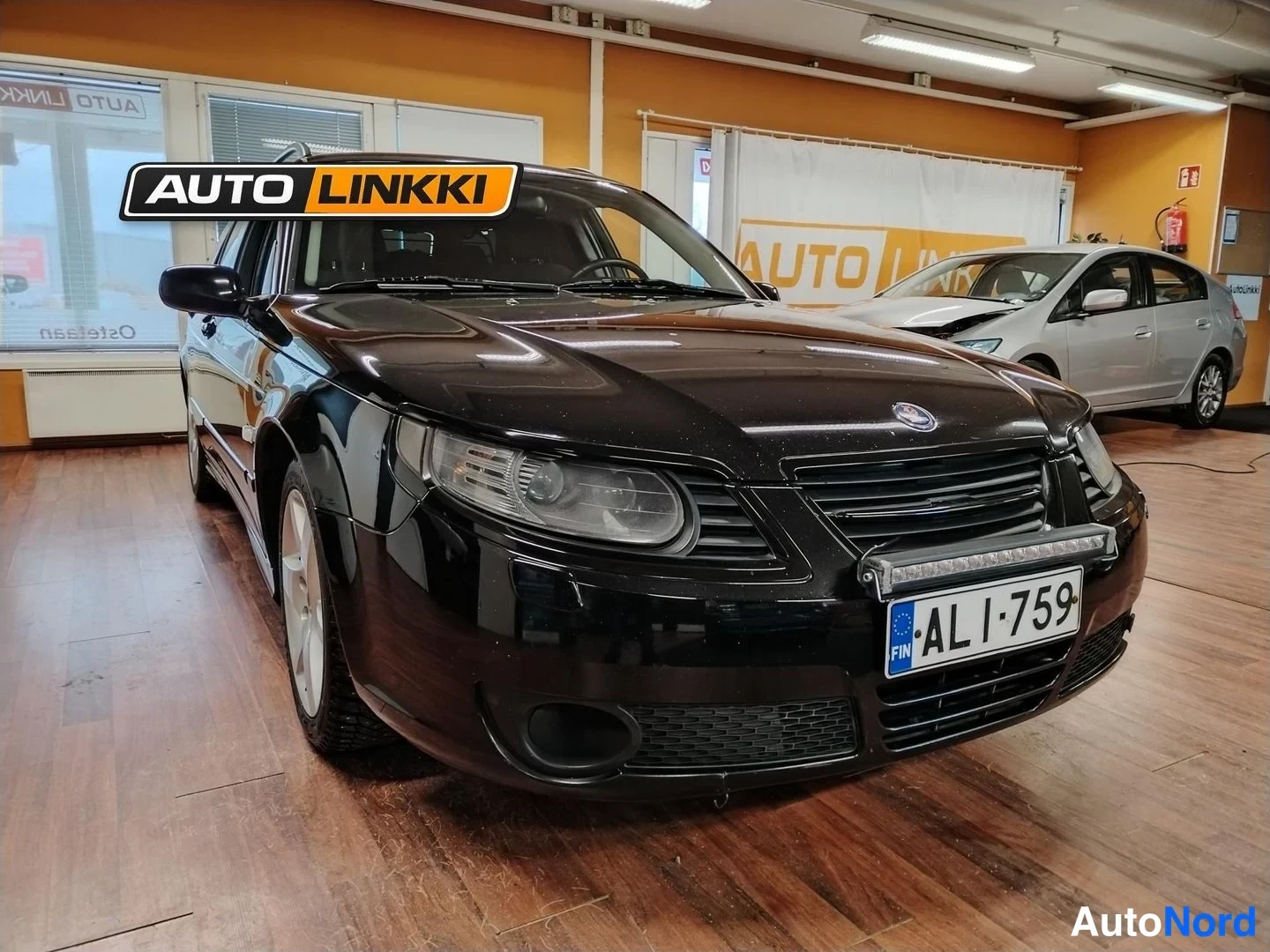 Etuviisto, nokka oikealle: Musta Saab 9-5 -farmari kuvattu etuviistöstä autosalissa, etupanssari ja rekisterikilpi näkyvissä.