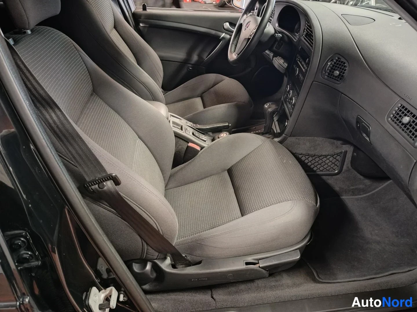 Edestä: Saab 9-5:n etupenkkien välikohtaus: kaksi tummanharmaaa kankaaverhoiltua istuinta, auton sisätilat mustat, ohjauspyörä ja keskikonsoli näkyvät.