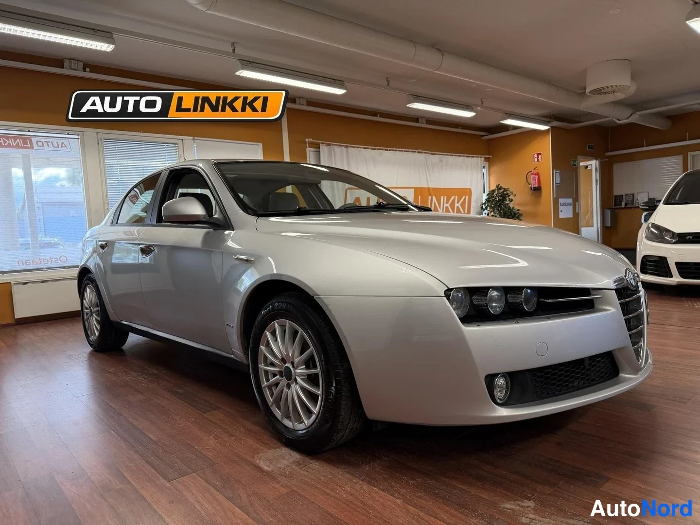 Etuviisto, nokka oikealle: Hopeanvärinen Alfa Romeo 159 -sedan nähty etukolmesta-auto kulmassa autonäyttelytilassa, keulan ja vasemman sivun näkyvissä.