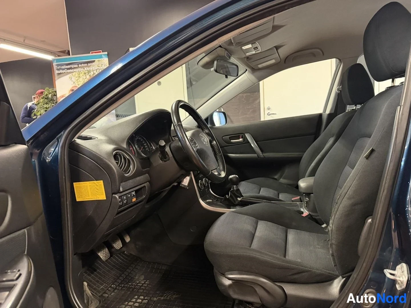 Muu: Auton etuistuin ja ohjaamo vasemman etuoven auki ollessa: Mazda-ohjauspyörä, analogiset mittarit, manuaalivaihteisto ja keltainen huoltotarra ovenkarmiin.