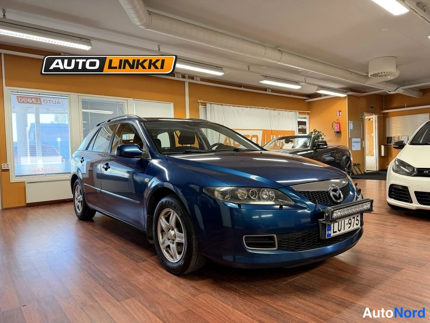 Etuviisto, nokka oikealle: Sininen Mazda 6 -farmari etukulmasta sisätiloissa, etupyörät ja konepelti näkyvät.