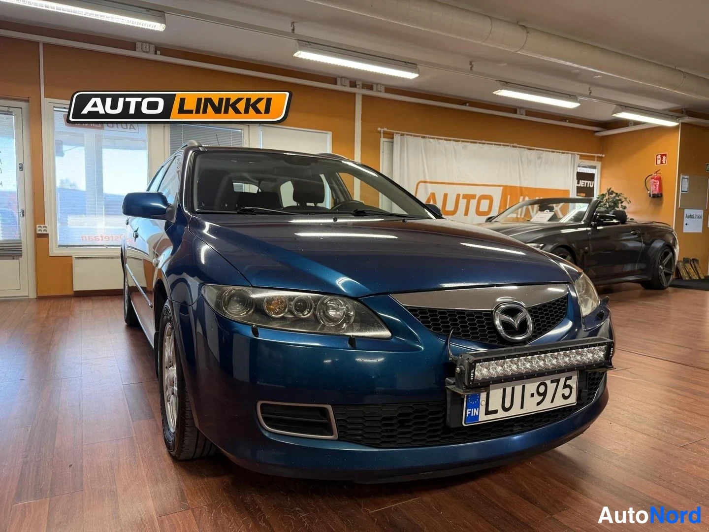Etuviisto, nokka oikealle: Sininen Mazda 6 -farmari etukulmasta sisätiloissa, etusäleikkö ja rekisterikilpi näkyvät.