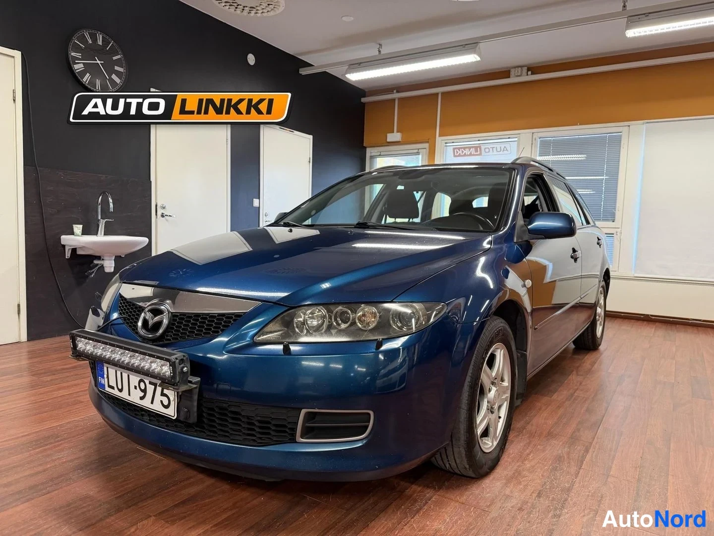 Etuviisto, nokka vasemmalle: Sininen Mazda 6 -farmari etukulmasta sisätiloissa, etusäleikkö ja ajovalot näkyvät.