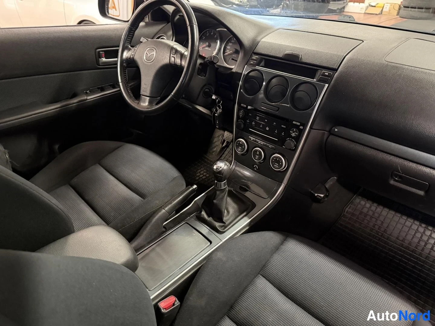 Muu: Auton ohjaamo vasemmalta katsottuna: Mazda-merkkinen ohjauspyörä, analogiset mittarit, manuaalivaihteisto ja keskikonsolin kelloradio.