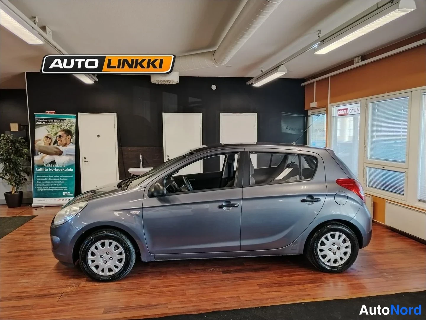 Etuviisto, nokka vasemmalle: Hyundai i20 -henkilöauto harmaana sivuprofiilista sisätiloissa, molemmat sivuovet näkyvissä.