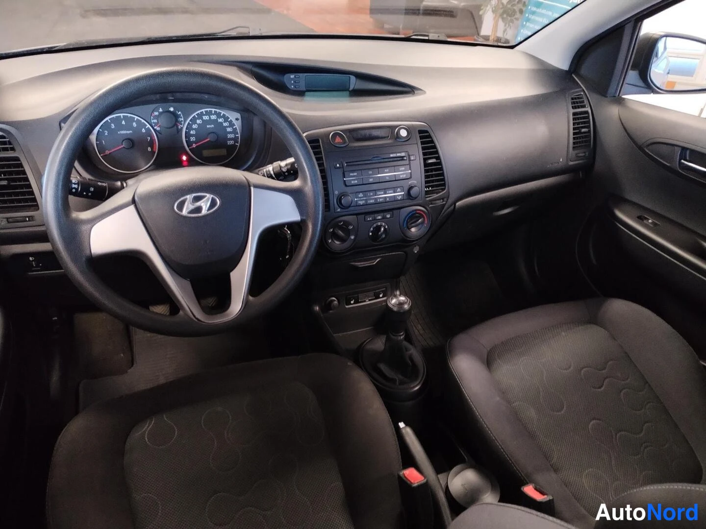 Muu: Kuljettajan näkökulma kojelautaan: Hyundai-ohjauspyörä, analogiset mittarit, radio ja manuaalivaihteiston vaihdankeppi.