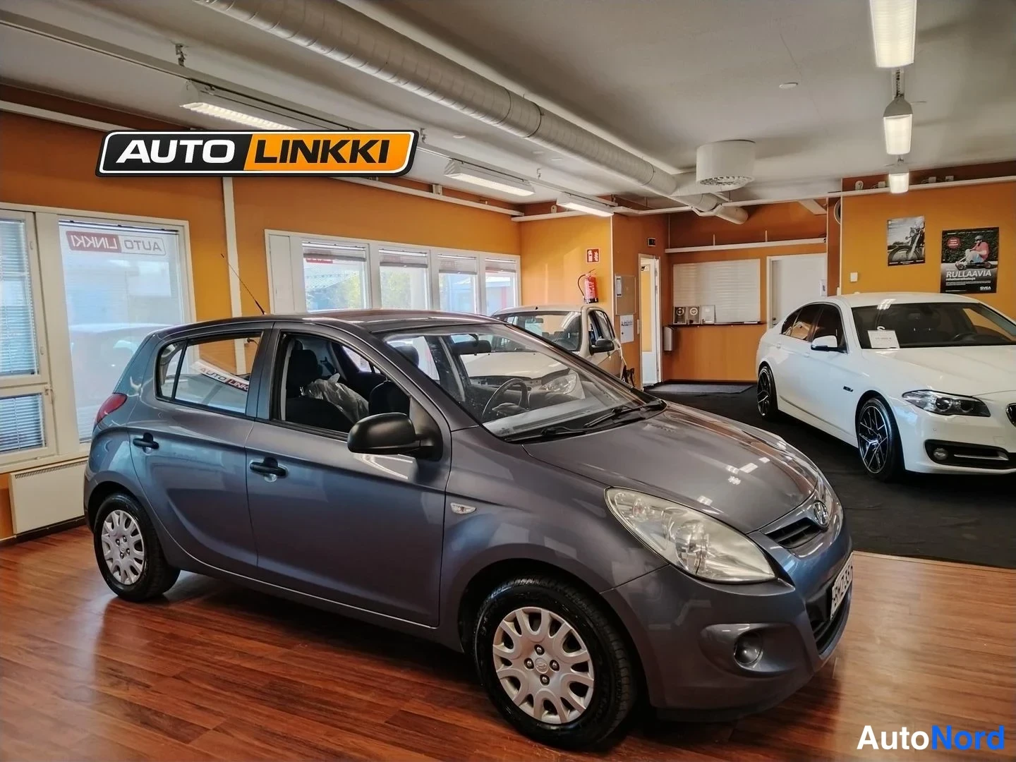 Etuviisto, nokka oikealle: Hyundai i20 -viistokuva etupuoli ja oikea kylki näkyvissä, auto tummanharmaa, sisätiloissa näyttelysalissa.