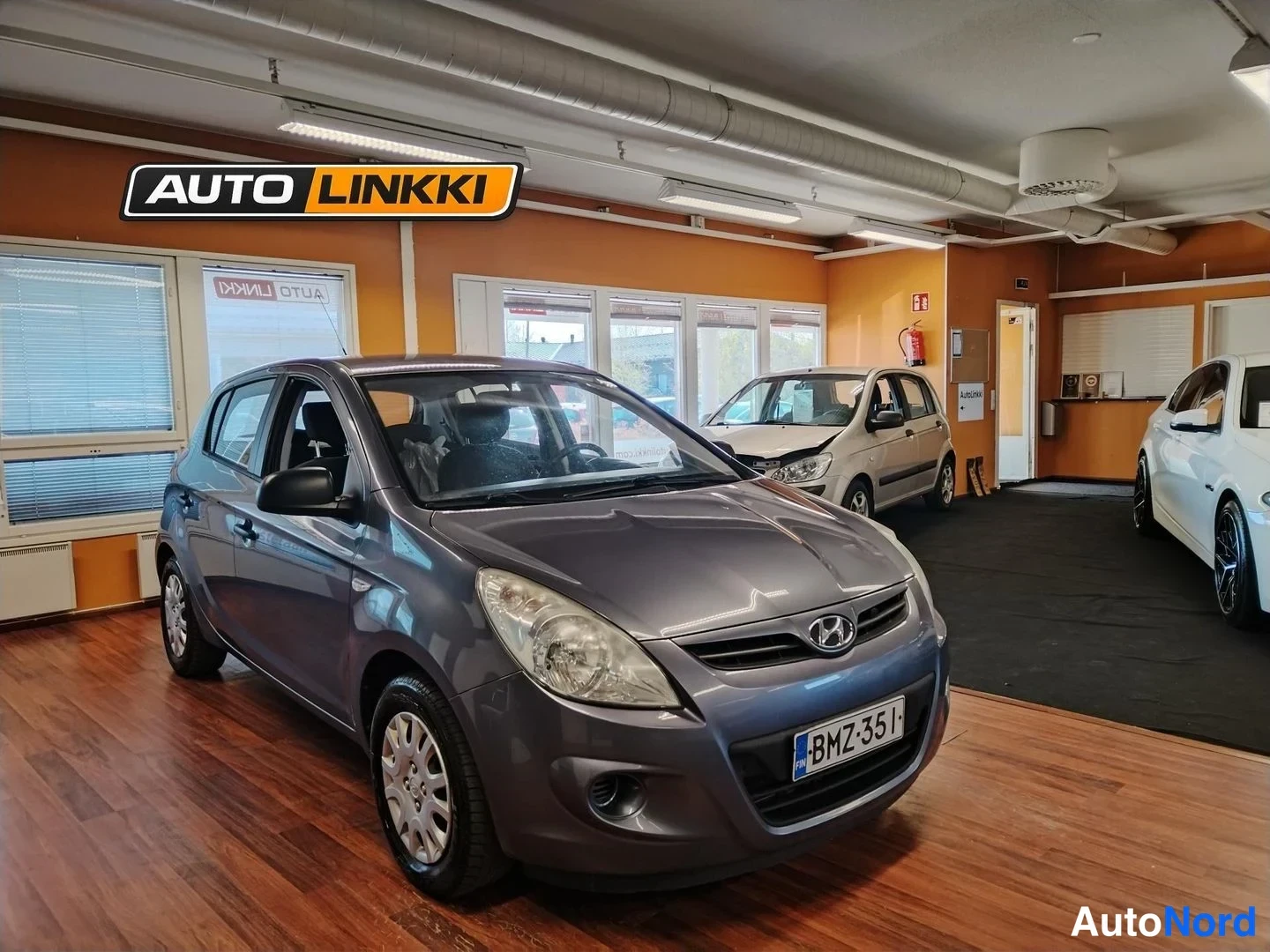 Etuviisto, nokka oikealle: Hyundai i20 etuviisto oikealta, rekisterikilpi, auto tummanharmaa näyttelysalissa.