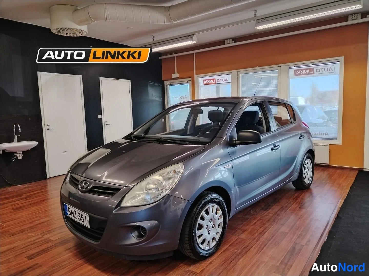 Etuviisto, nokka oikealle: Hyundai i20 etuviisto vasemmalta, rekisterikilpi, auto tummanharmaa sisätiloissa.