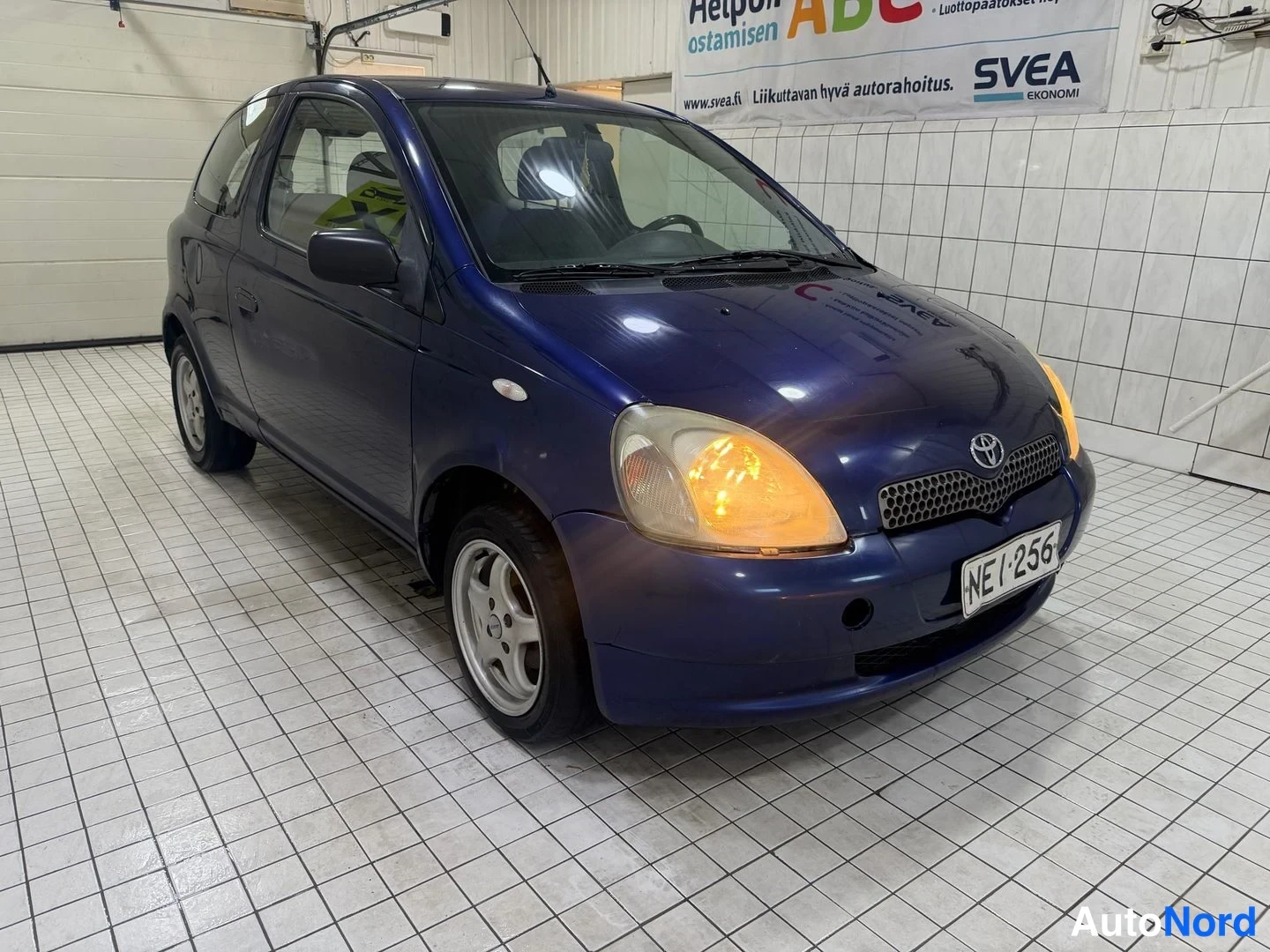 Etuviisto, nokka oikealle: Sininen kolmiovinen hatchback etukulmasta oikealta kuvattuna, etuvalot päällä, rekisterikilpi, hopeavanteet.