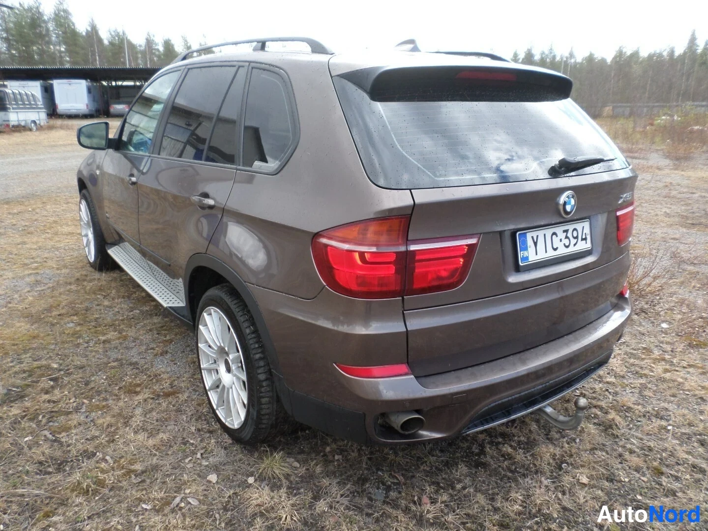 Takaviisto: BMW X5:n takaosa ja vasen kylki kolmesta neljäsosasta, ruskeaa pintaa, rekisteritunnus, perävaske ja trailerikoukku näkyvissä.