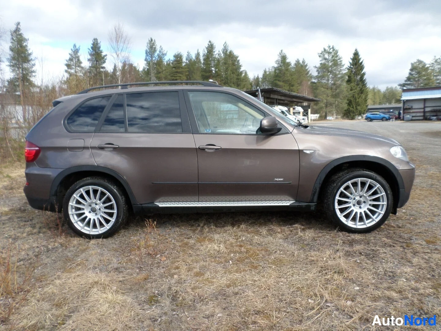 Sivulta: BMW X5:n vasen sivuprofiili avoimella paikalla, ruskea kori, kattokaiteet ja hopeiset sivuluskat näkyvissä.