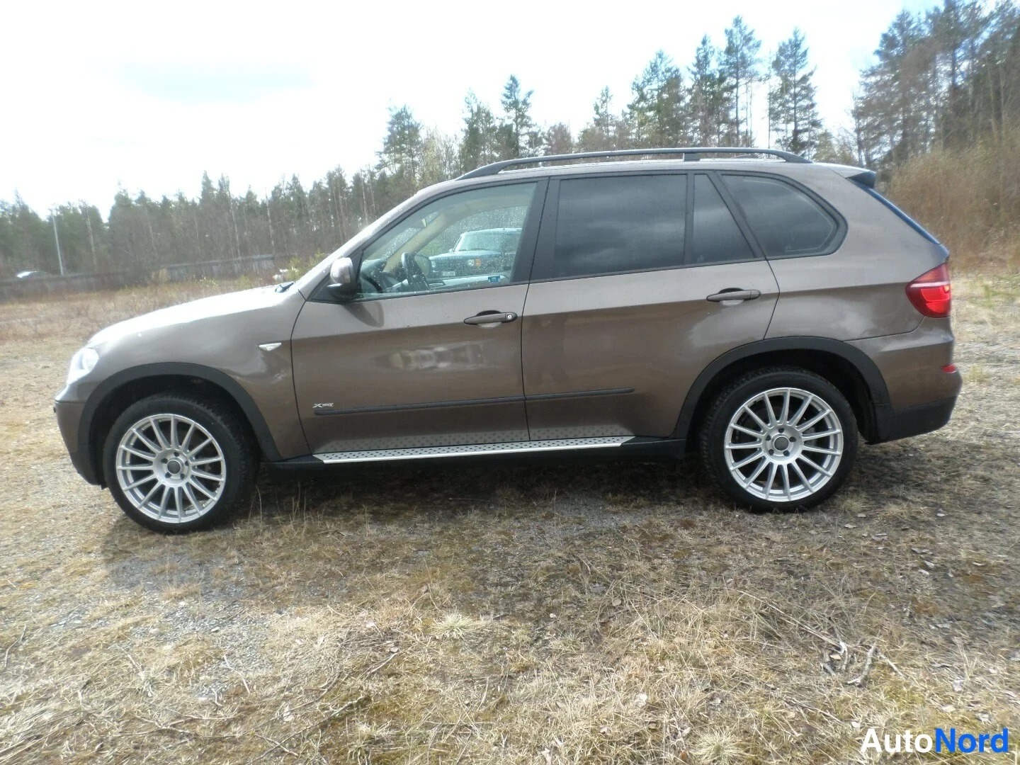 Sivulta: BMW X5:n vasen sivuprofiili, ruskea kori, kattokaiteet, sivuluskat ja hopeiset kevytmetallivanteet näkyvissä.