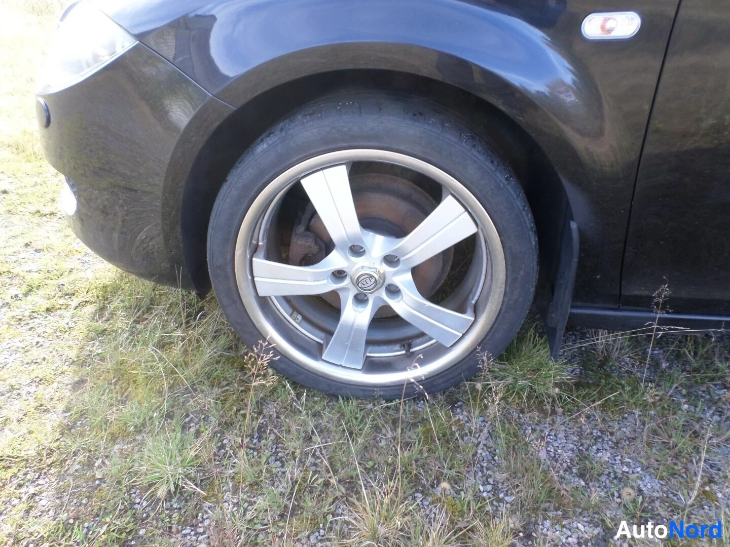 Etuviisto, nokka vasemmalle: Mustan auton etupyörän lähikuva, hopea viisipuolainen kevytmetallivanne.