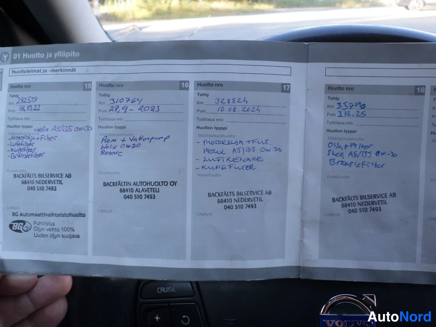 Asiakirja: Close-up of a service book (service log) opened to multiple pages showing maintenance records. The steering wheel in the background shows a cruise control butto