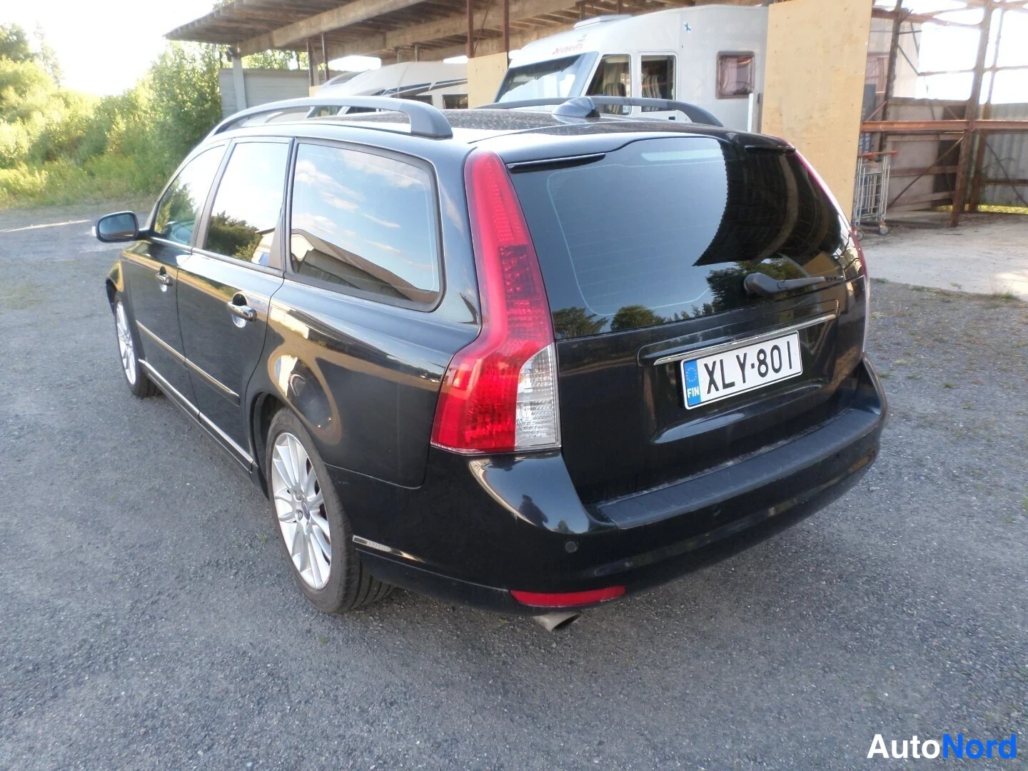 Takaviisto: Musta Volvo V50 -maasturimalli seisoo asfaltilla, näkyvissä auton takaosa ja vasen kylki. Takavalot ovat punaiset ja rekisterinumero on. Katossa on katotelineet