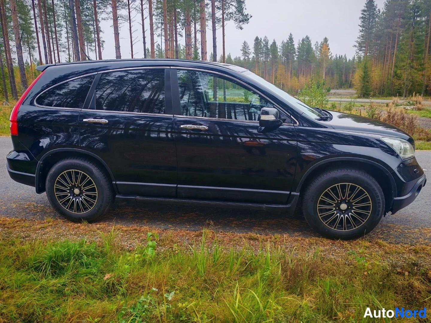 Sivulta: Musta Honda CR-V täysin profiilikuvassa seisomassa metsäautotien reunalla, molemmat pyörät ja metalliset vanteet näkyvät