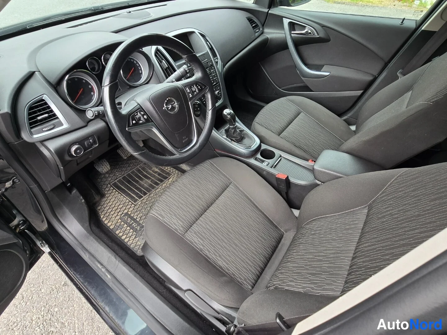 Edestä: Opel Astra etusumu, musta kangasverhoilu, ASTRAlla varustettu kumimatto ja manuaalivaihteisto.
