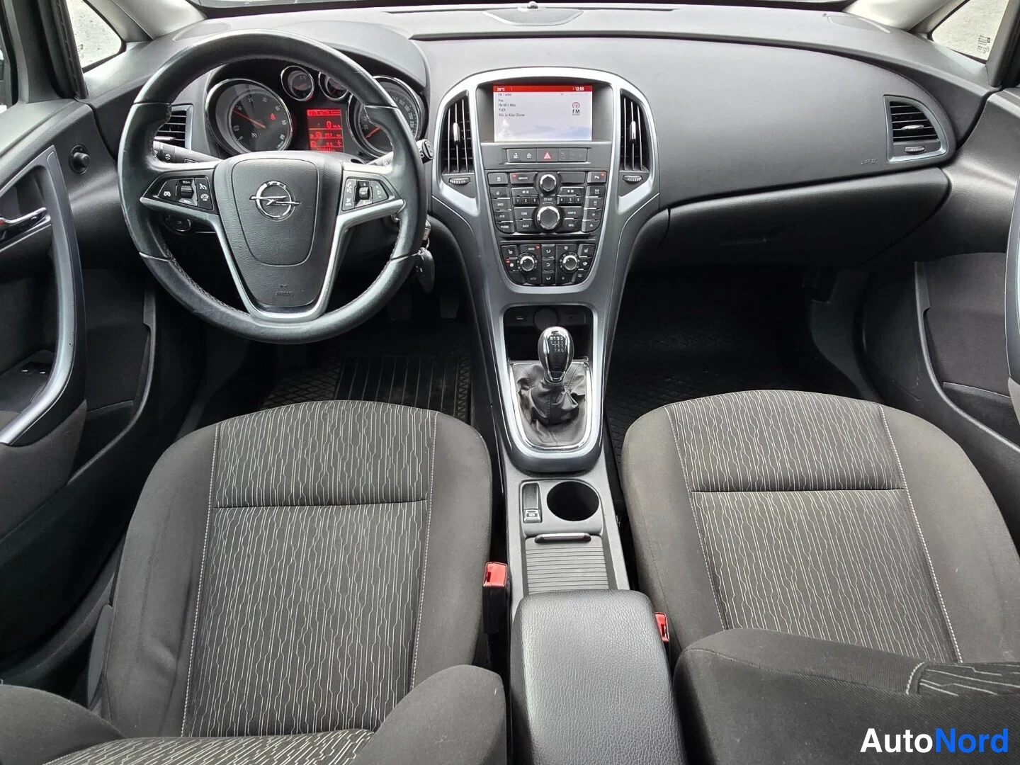 Edestä: Opel Astra kojelauta ja manuaalivaihteisto, näyttö päällä, kaksi etuistuinta.