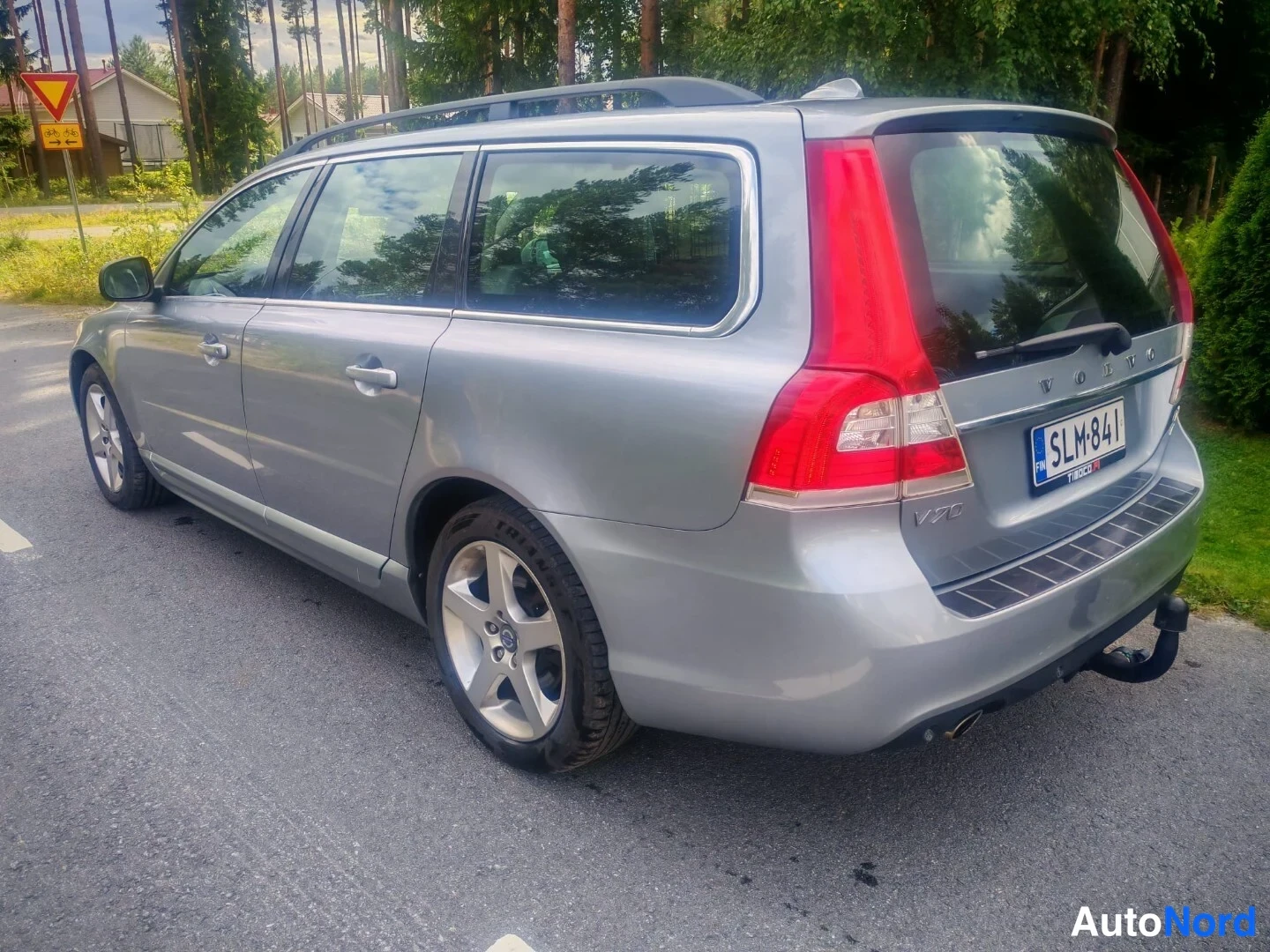 Takaviisto: Hopeanharmaa V70-asema-auto takaa vasemmalta kolmanneltasektorilta, rekisterikilpi, kattotankot, takavalo, lukkokeko takaossa, tie ja vihreys taustalla.