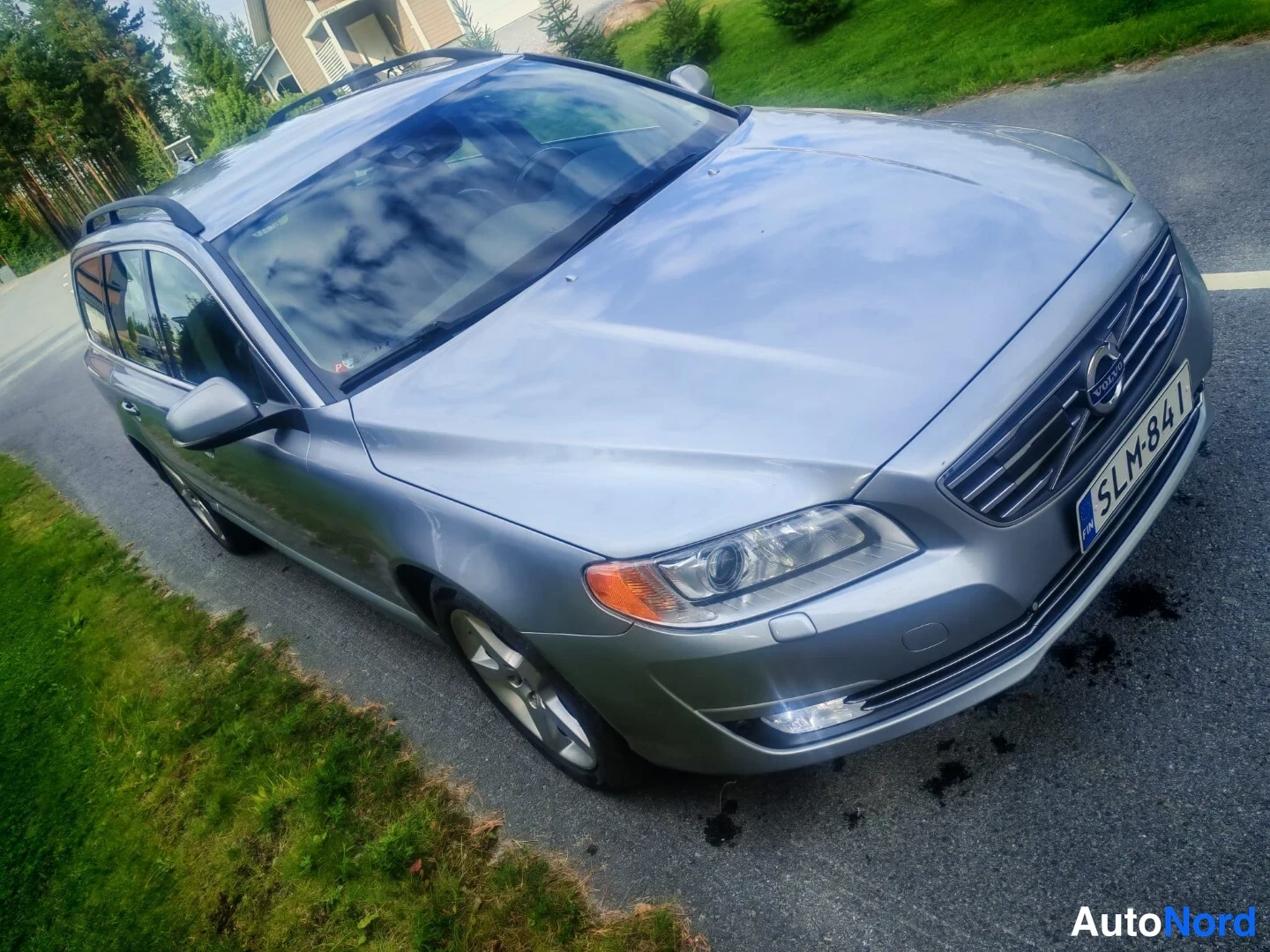 Etuviisto, nokka oikealle: Kuvan oikea yläkulma: harmaa Volvo-asema-auto, jossa näkyy etuosa ja vasen sivu. Etukalustossa on Volvo-logo ja rekisterikilpi. Kattoon on asennettu kato