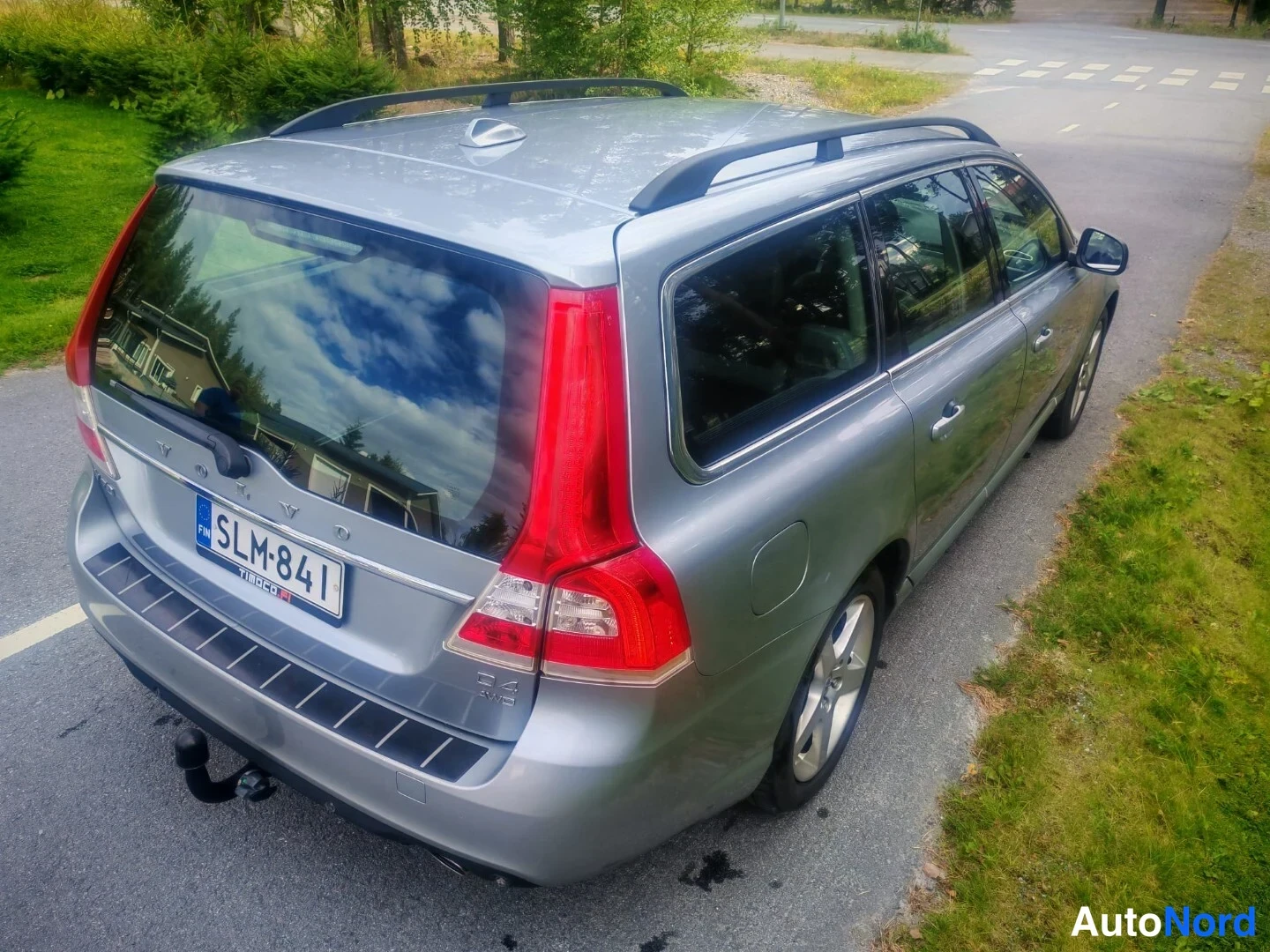 Takaviisto: A silver Volvo V70 station wagon parked on an asphalt road next to green grass, featuring roof rails, rear tow bar, and vertical tail lights