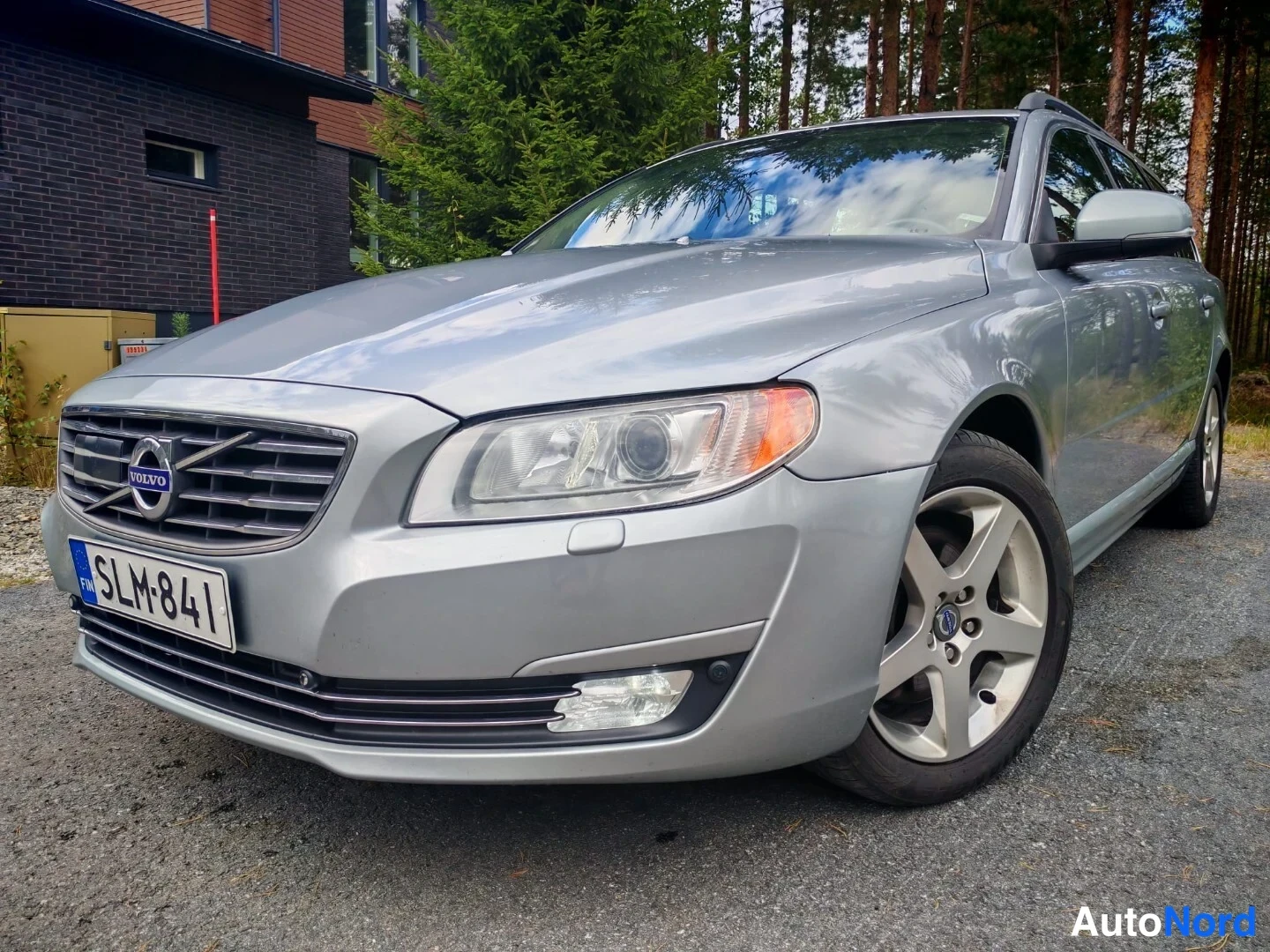 Etuviisto, nokka vasemmalle: Hopeanharmaa Volvo V70 -asema-auto kuvattu kolmanneltasektorilta edestä vasemmalta, rekisterikilpi, hiekkapäällysteinen piha, rakennus ja metsä taustal