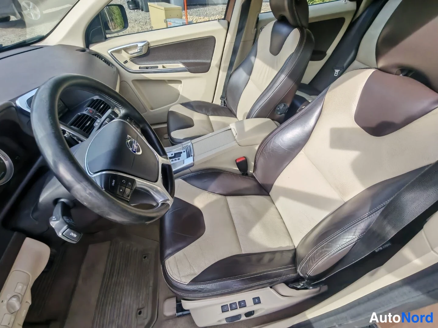 Muu: Ohjaamon näkymä kuljettajan puolelta: Volvo-ohjauspyörä, tumma kojelauta ja beige-ruskeat nahkaistuimet.