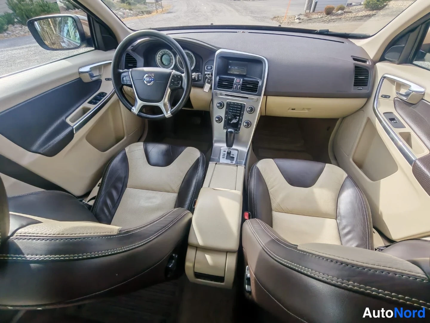 Ohjaamo: Interior view of a car showing front seats with beige and dark brown leather upholstery, dashboard with two-tone trim (beige and black), steering wheel with con