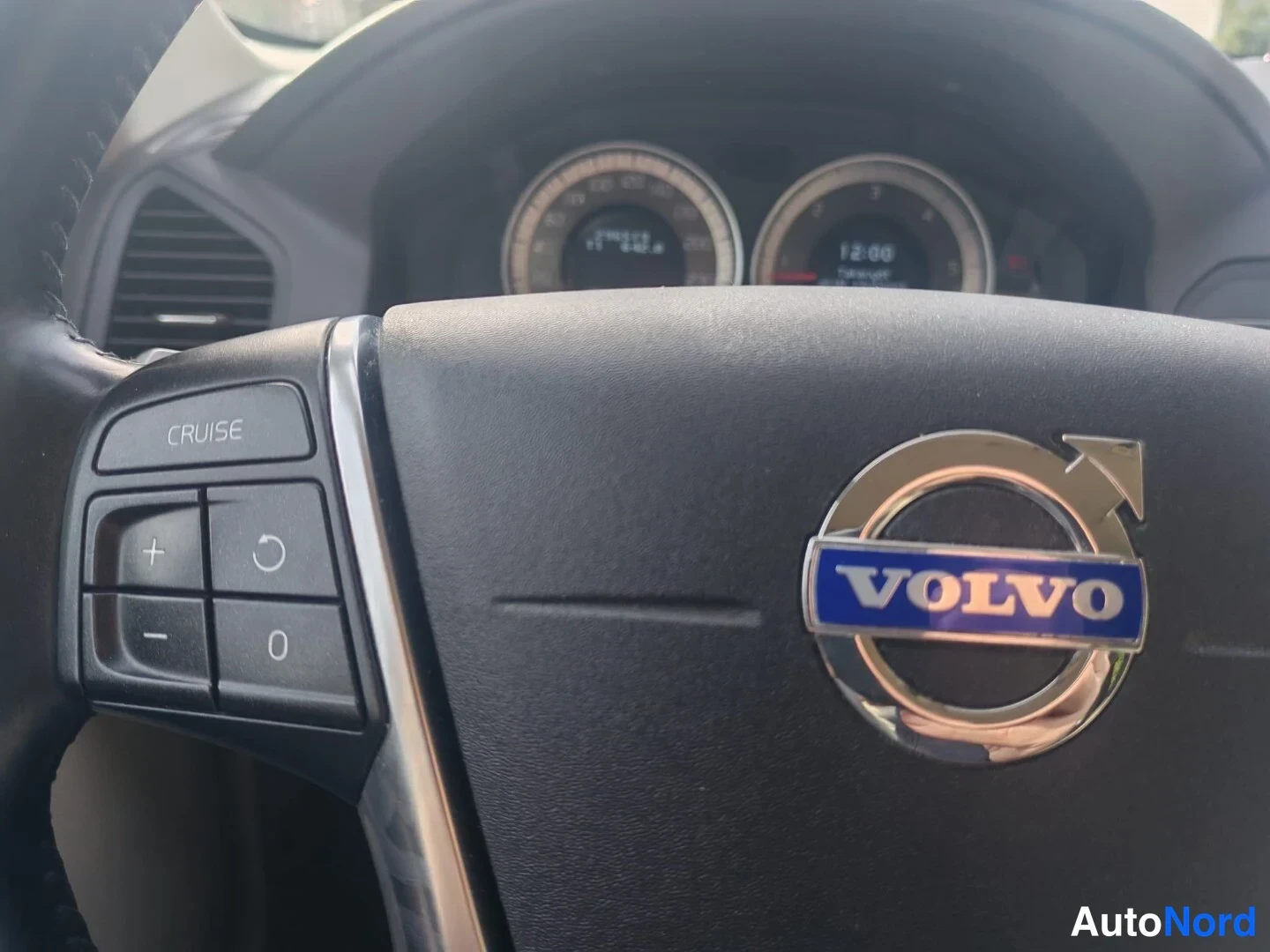 Muu: Volvo-ohjauspyörän keskusta lähikuva: VOLVO-emblem, CRUISE-näppäimet vasemmalla ja mittaristo taustalla näkyvällä kellonajalla 12:00.