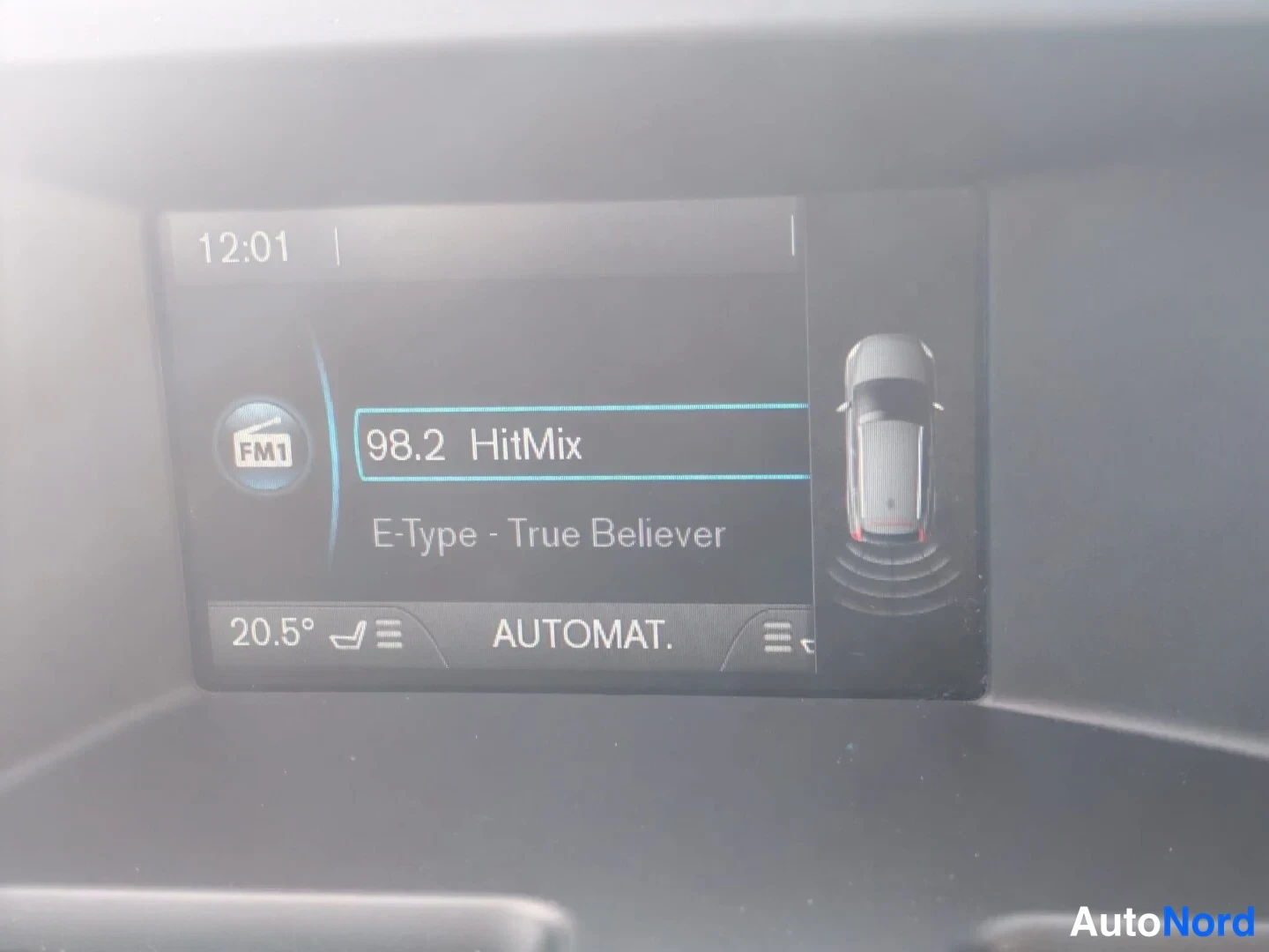 Muu: Volvo XC60:n kojetaulun näyttö: radio FM1 98.2 HitMix (E-Type - True Believer), kellonaika 12:01 ja parkkitutkagrafiikka takana