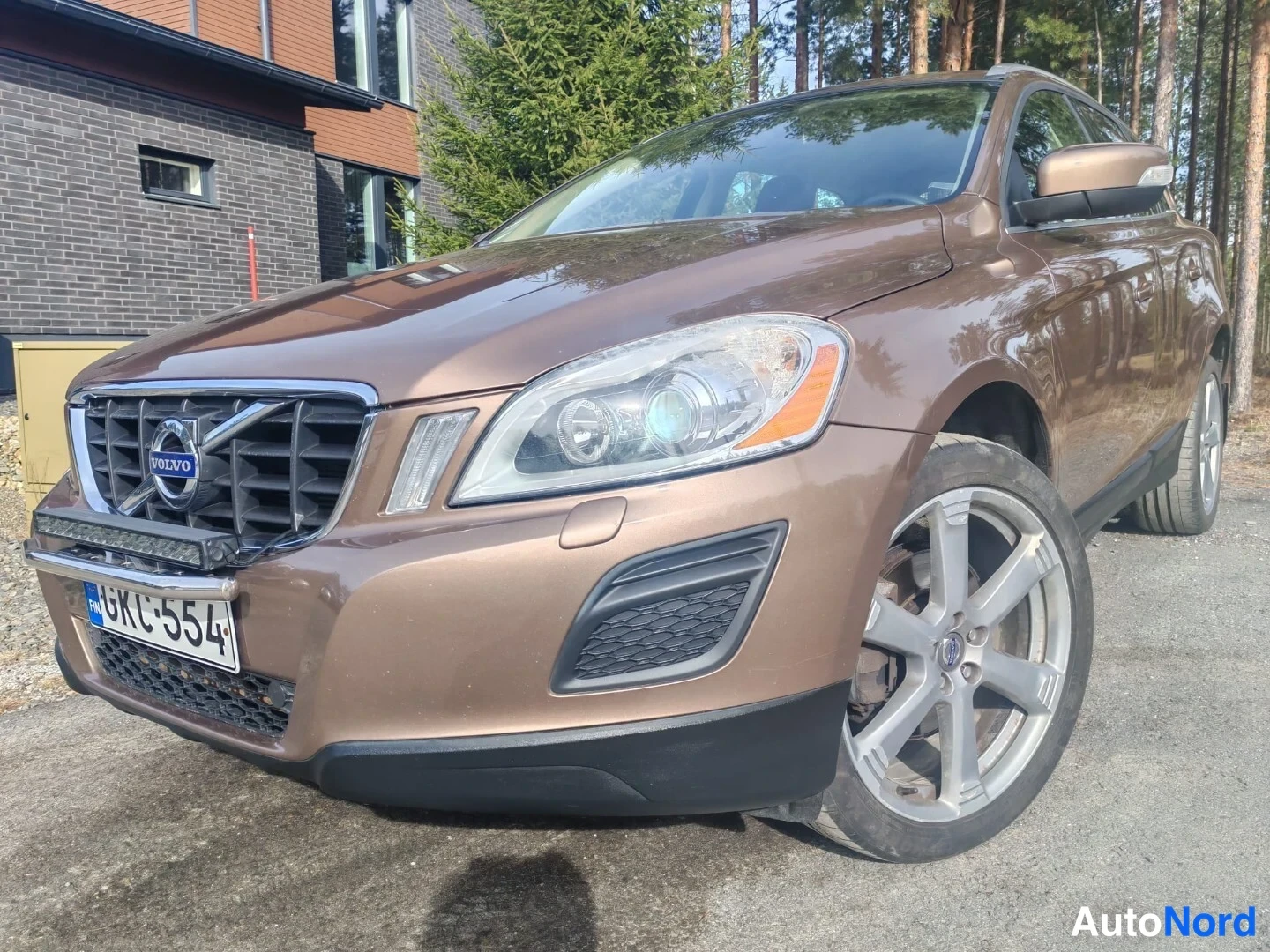 Etuviisto, nokka vasemmalle: Ruskea Volvo XC60 kulmassa edestä vasemmalta, etusäleikkö, ajovalot ja hopeiset vanteet näkyvissä, rekisterikilpi.