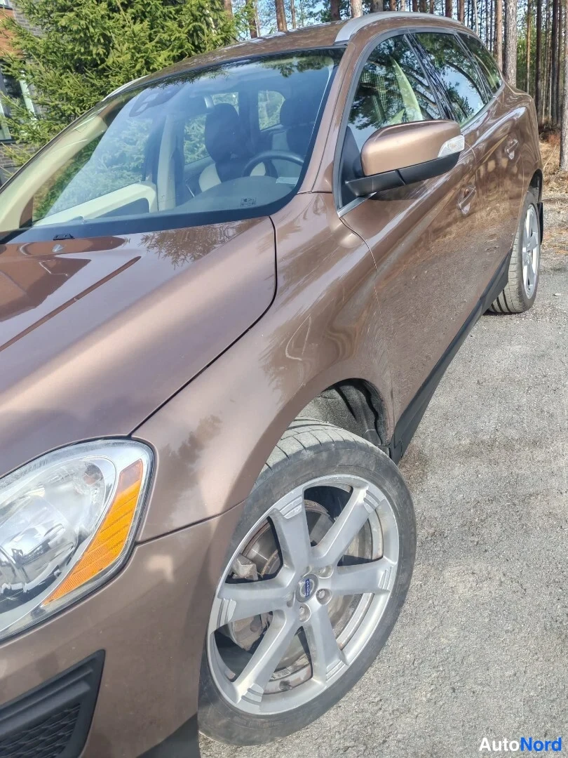 Etuviisto, nokka vasemmalle: Ruskean Volvo XC60:n etusarvi ja etupyörä lähikuvassa, tuulilasi ja sivupeili näkyvissä.