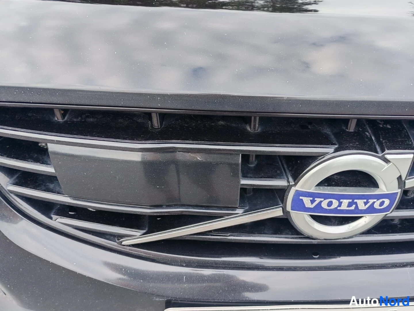 Yksityiskohta: Lähikuva Volvon logosta etusuojuksessa. Logossa on teksti 'VOLVO' sinisellä pohjalla ja hopeisella renkaalla ympäröitynä. Etusuojuksen ruudut ja metallipallit o