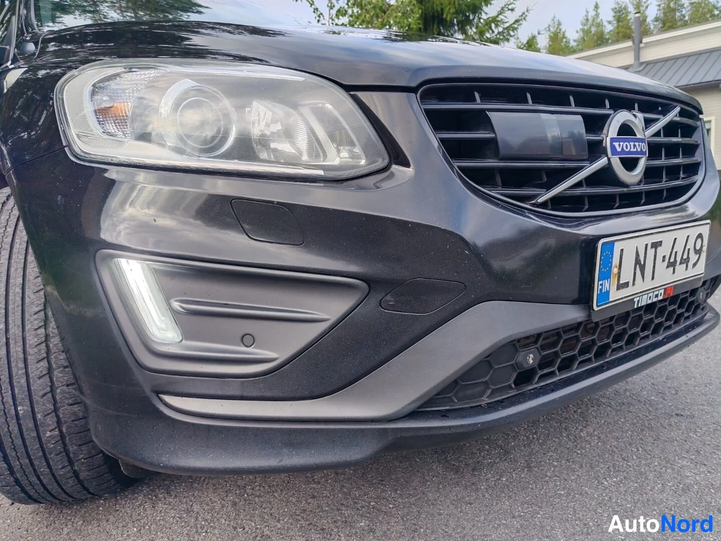 Edestä: Front fascia detail showing Volvo grille with logo, left headlight and fog light assembly on a black car.