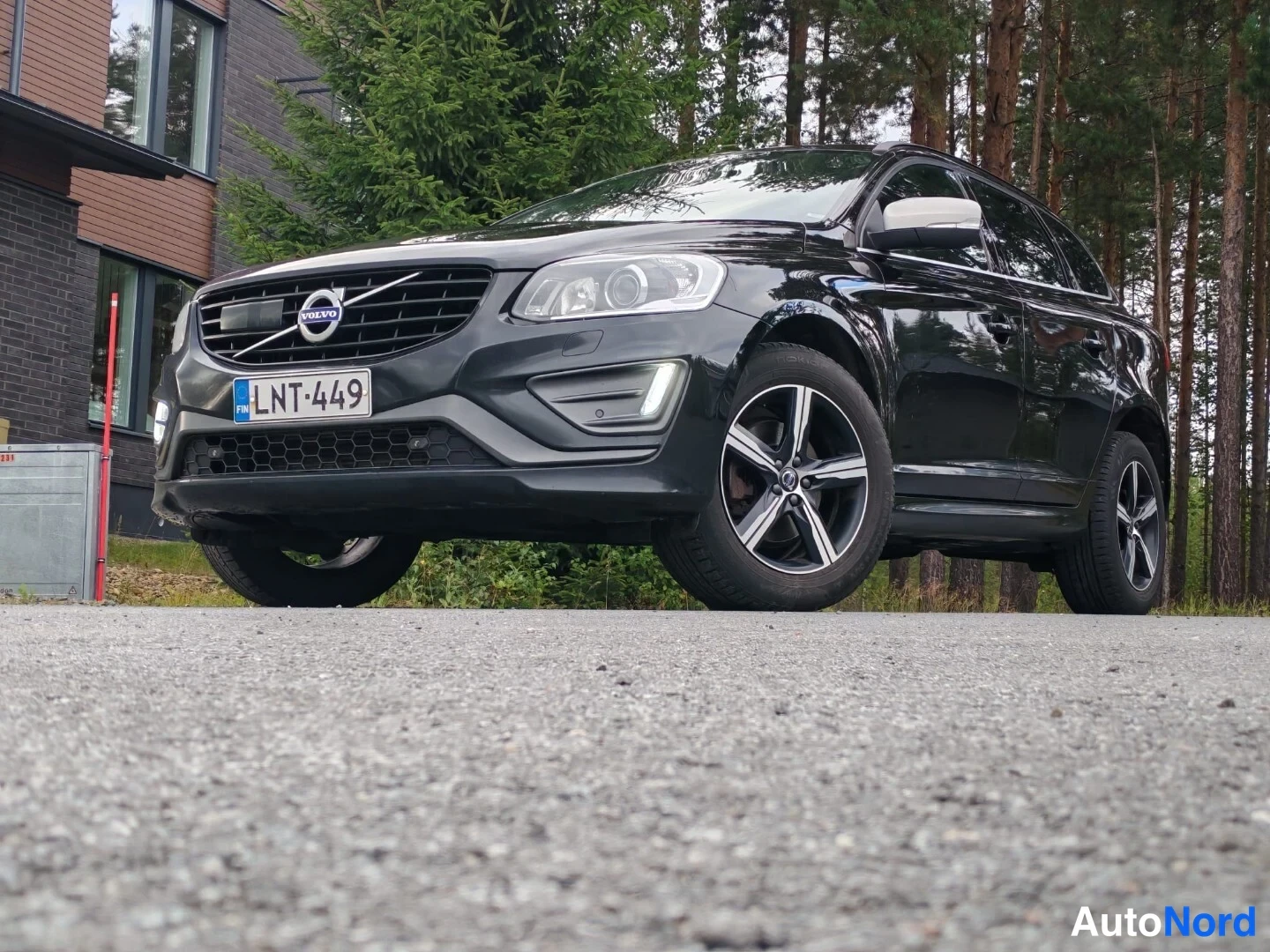 Etuviisto, nokka vasemmalle: Musta Volvo XC60 kolmasosakuvana edestä vasemmalta asfaltilla, talo ja metsä taustalla; rekisterikilpi.
