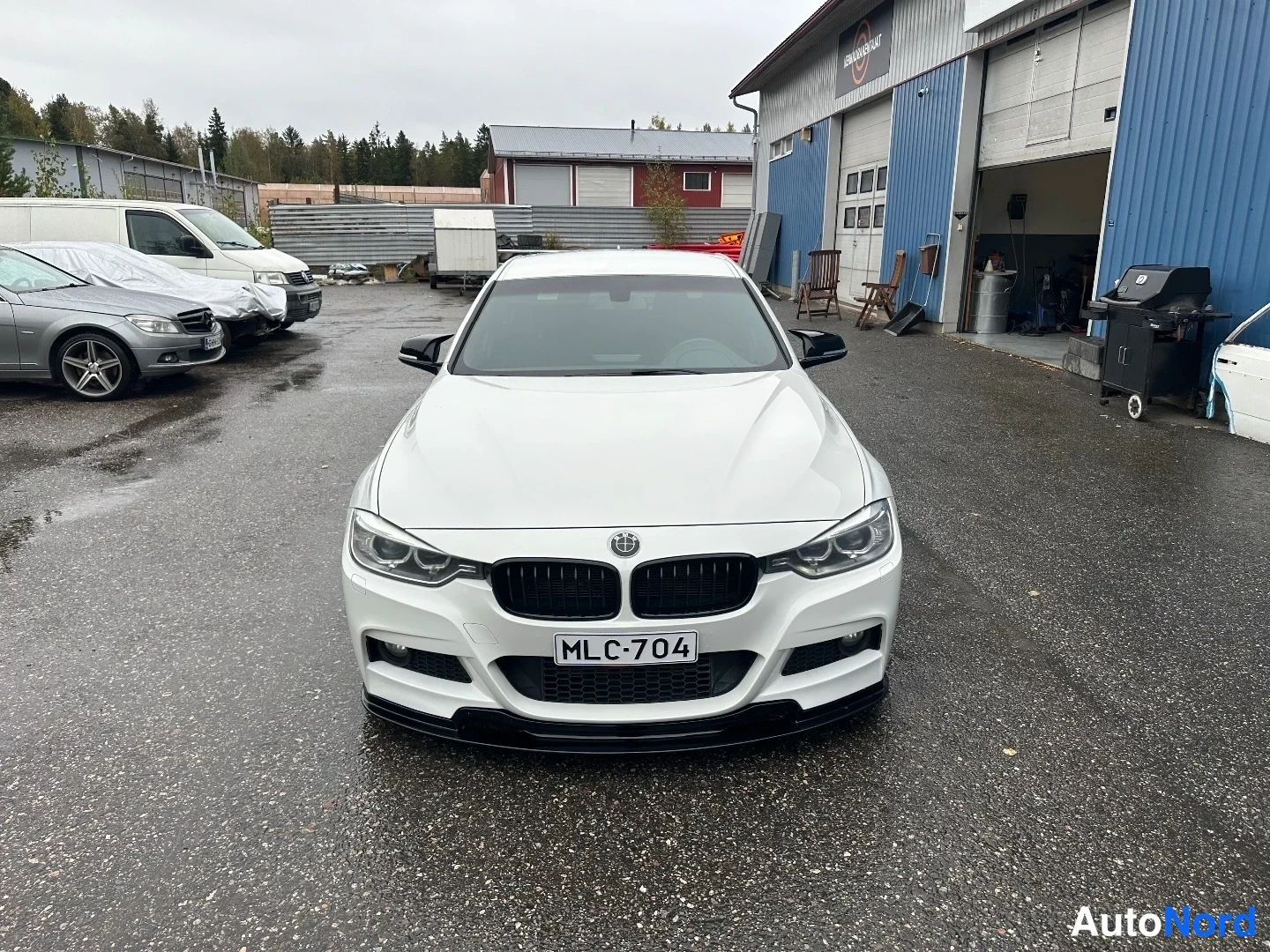 Edestä: Valkoinen BMW-sedan suoraan edestä päin kuvattuna pihalla, rekisteritunnus.