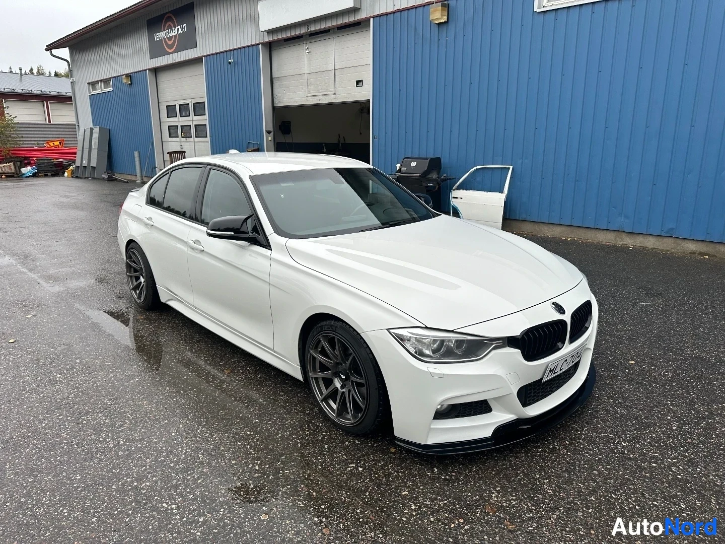 Etuviisto, nokka oikealle: Valkoinen BMW-sedan etukulma oikealta puolella kuvattuna korjaamon pihalla, tummat kevytmetallivanteet.