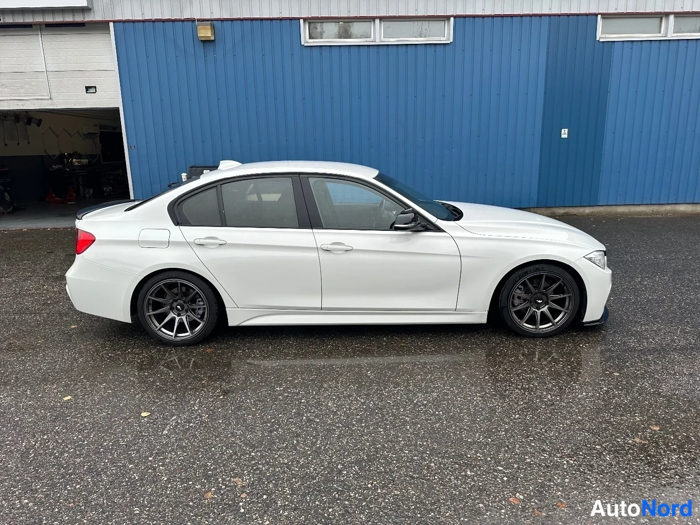 Sivulta: Valkoisen BMW-sedanin sivukuva sinisen rakennuksen edessä; tummat kevytmetallivanteet näkyvissä.