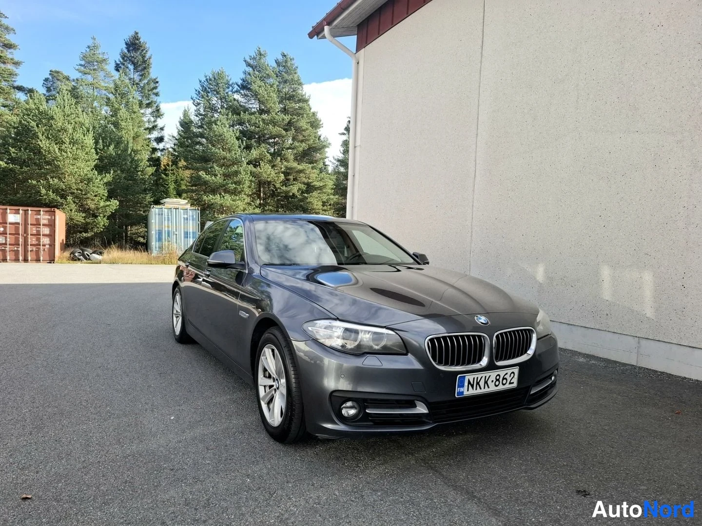 Etuviisto, nokka oikealle: Tummanharmaa BMW-sedan kolmenvartalonäkymä edestä oikealta, rekisteritunnus, parkissa valkoisen hallin edessä.