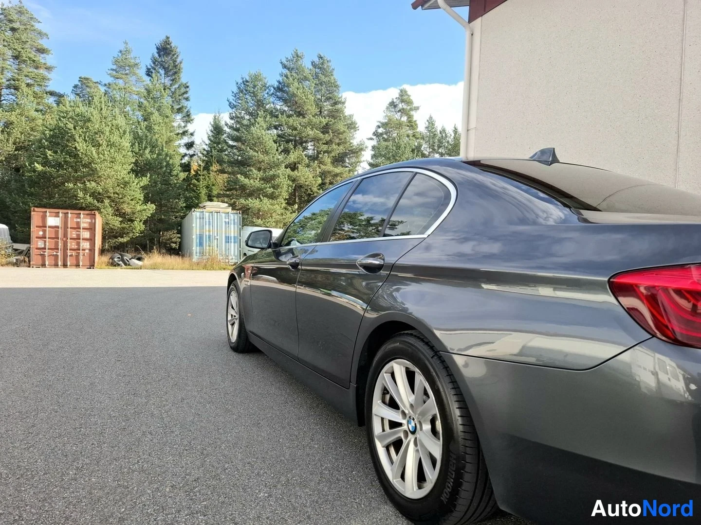 Takaviisto: Tummanharmaa BMW-sedan takakolmivartalokuvakulmasta, takaosa ja vasen sivu näkyvällä pihalla.