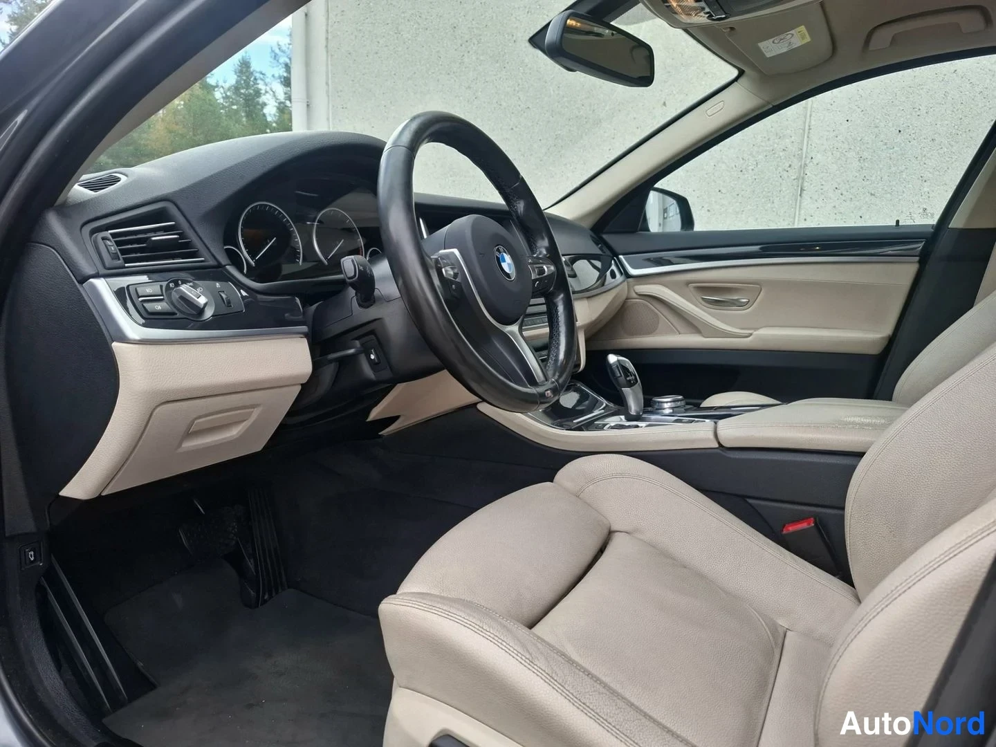 Sivulta: BMW:n sisätilat ohjaamosta kuvattuna: beige nahkapenkit, musta ohjauspyörä BMW-tunnuksella, instrumentit ja automaattivaihteen valitsin.