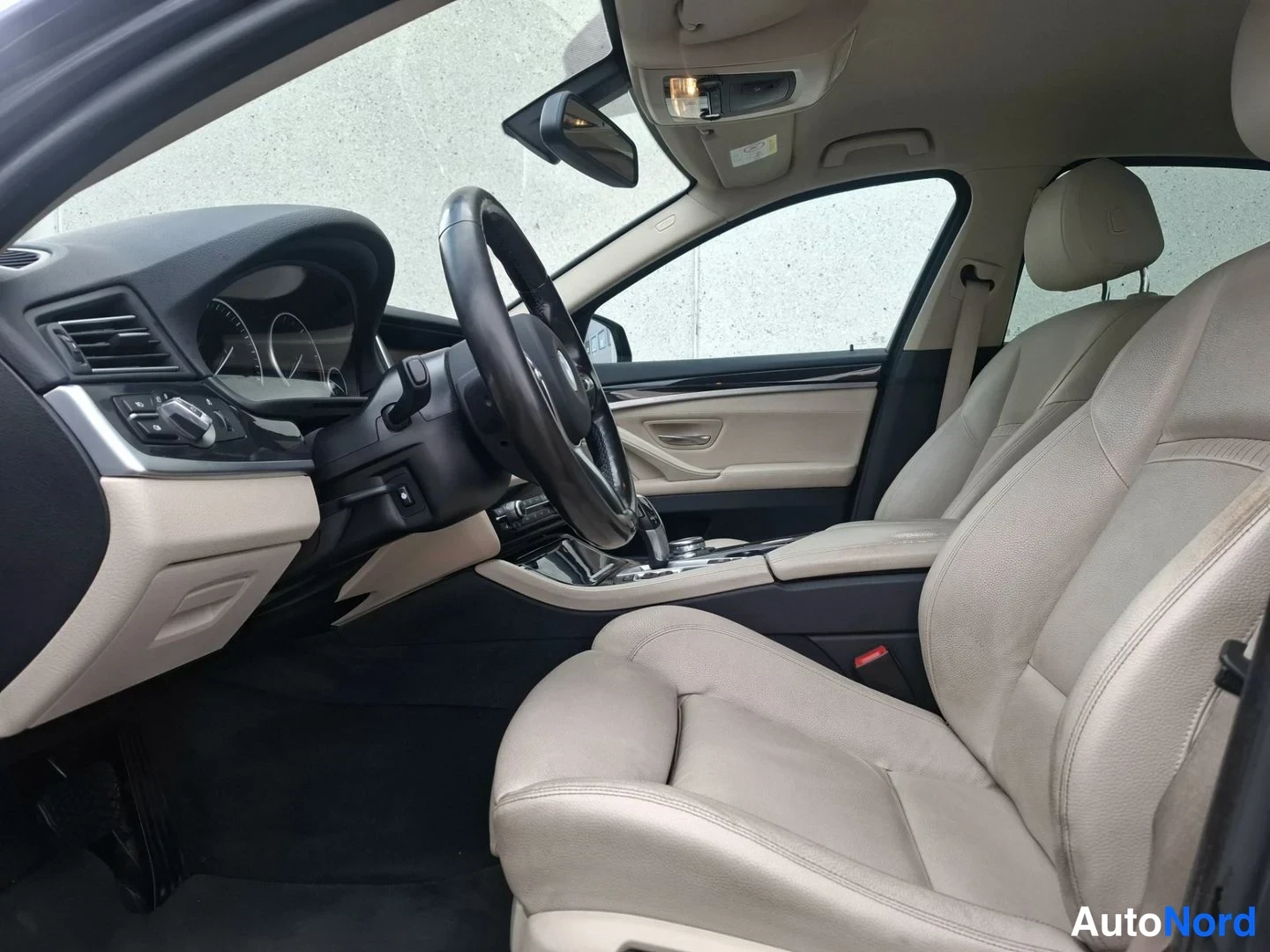Ohjaamo: Interior shot showing beige leather front passenger seat and dashboard from the rear door. A multifunction steering wheel and center console gear selector are v