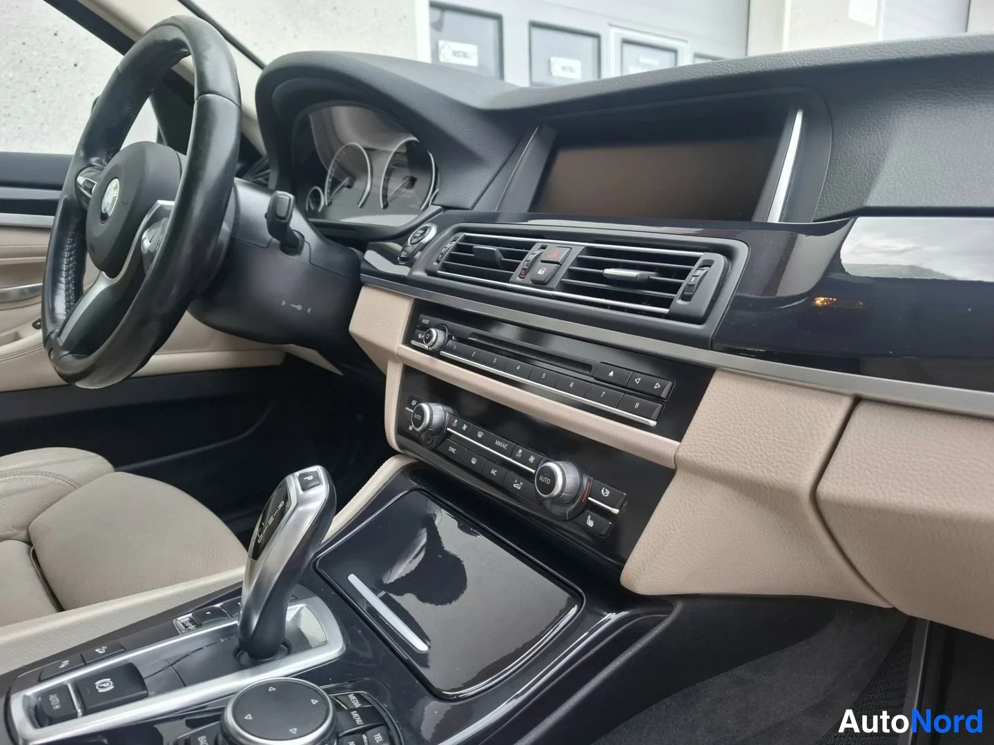 Yksityiskohta: Close-up of the center console. An automatic gear shifter is positioned next to a rotary dial controller and USB/Auxiliary input ports. The infotainment screen
