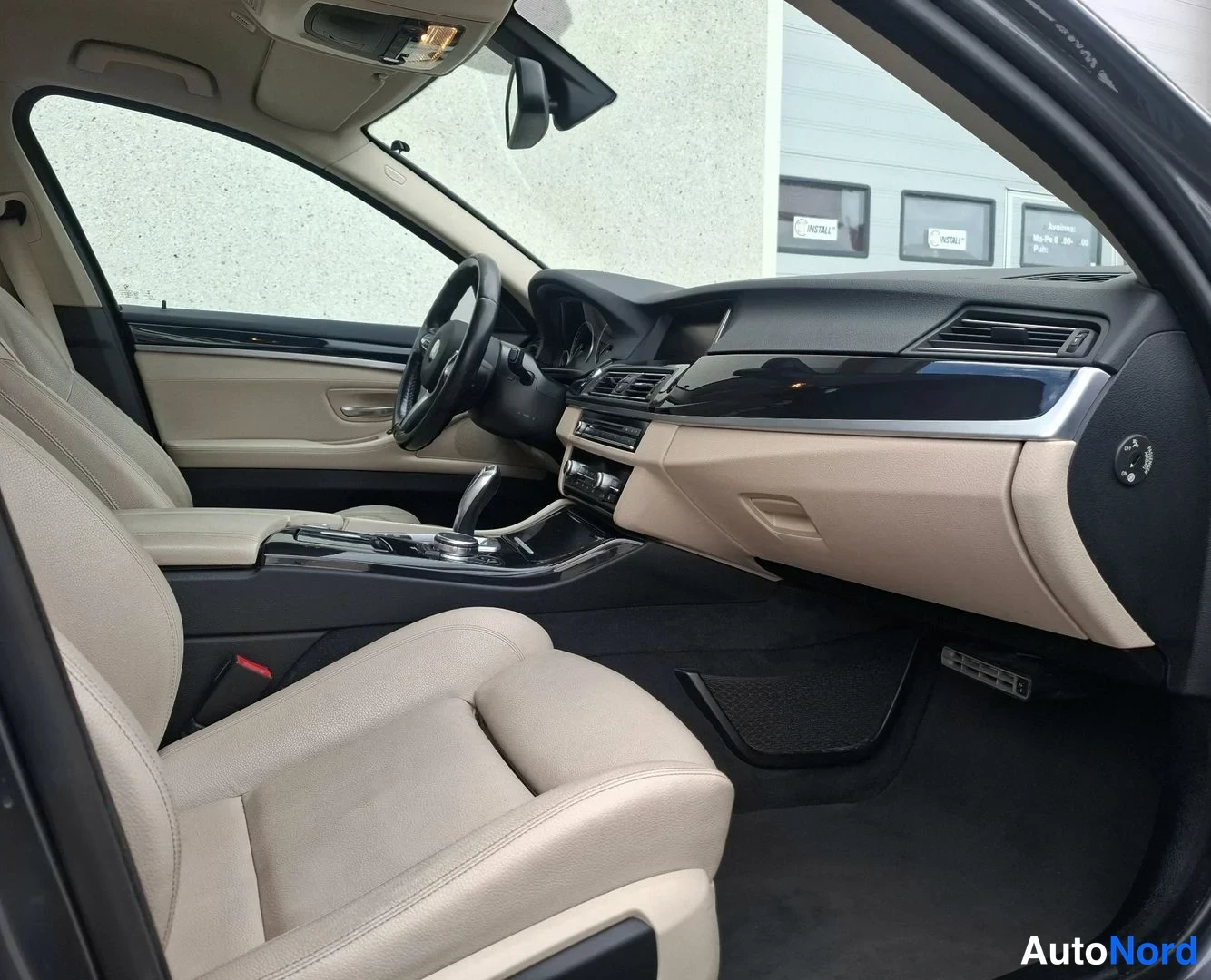 Ohjaamo: Wide interior view looking into the cabin from the open driver's side door. It shows beige leather bucket seats, the steering wheel, and two-tone dashboard trim