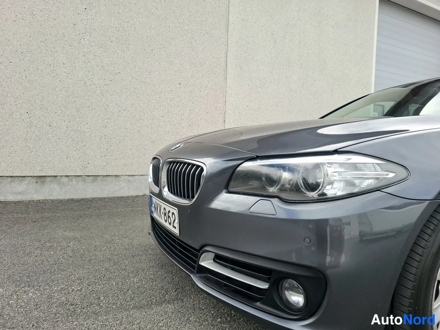 Edestä: Auton etuosa lähikuvassa: BMW-merkki etupanssarissa, etuvalo ja sumuvalot tummassa etupuskurissa. Rekisterikilpi.